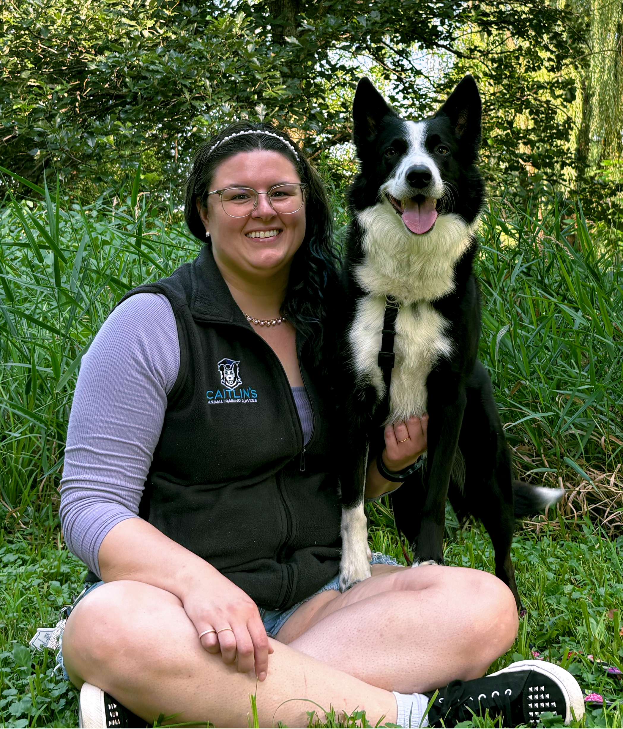 A woman with blue hair sitting in the grass with a black and white dog perching on her leg